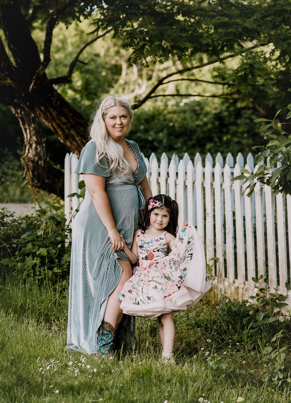 A pregnant woman and a little girl are standing next to each other in front of a white picket fence.
