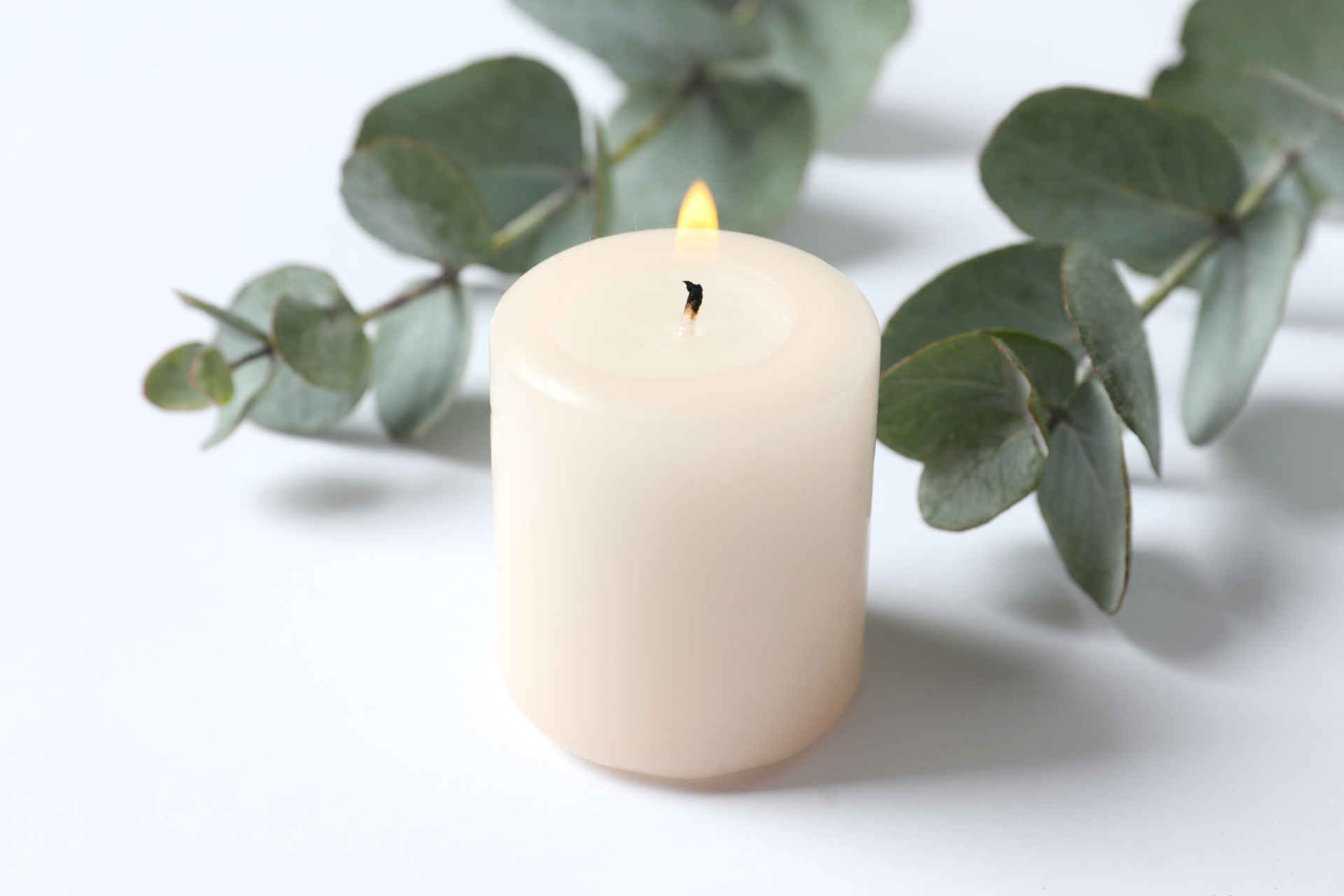 Lit cream-colored candle with a sprig of eucalyptus on a white surface.