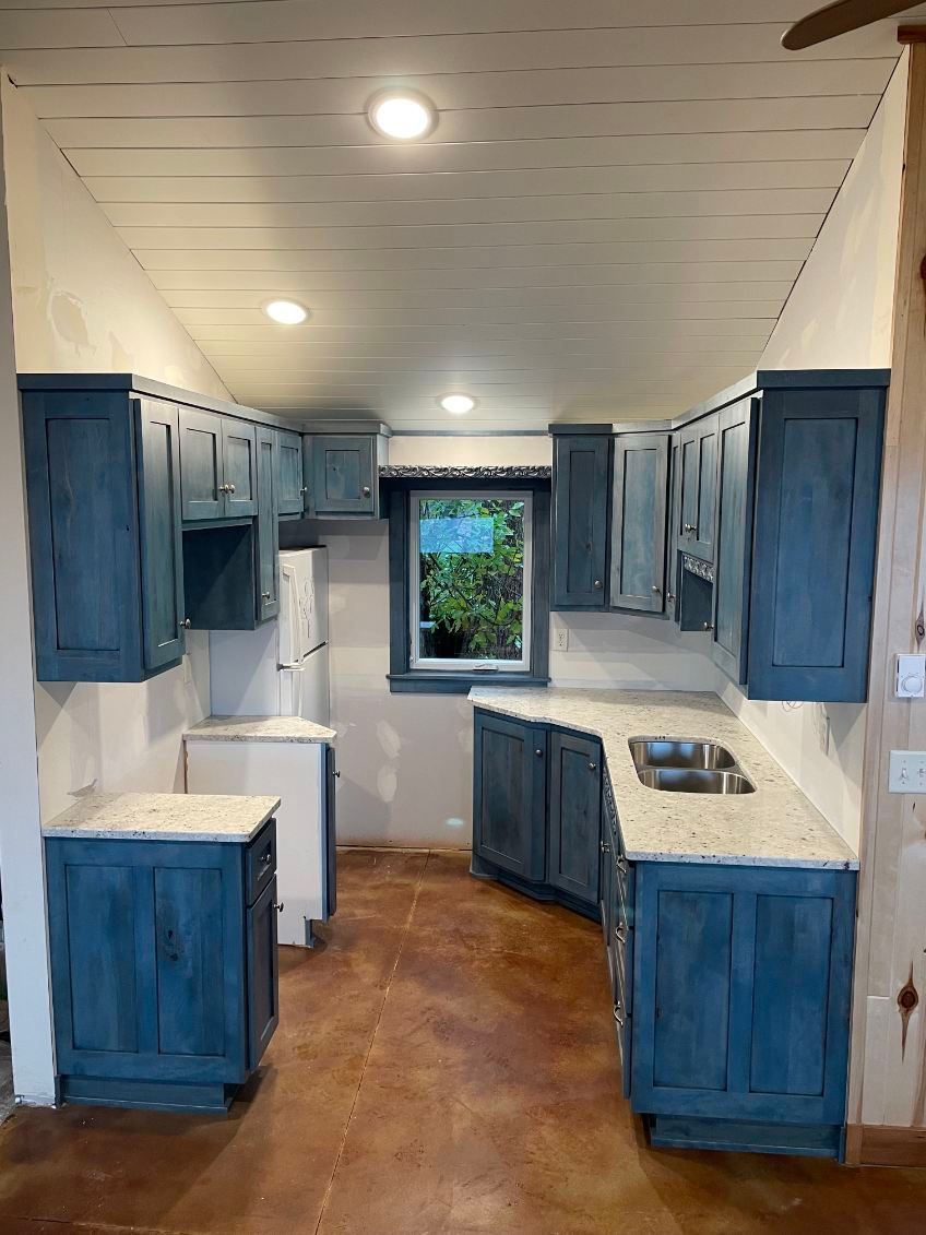 Blue cabinets in a small kitchen with a window; light-colored countertops and flooring.
