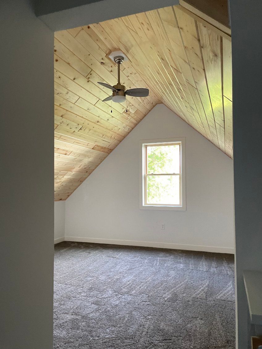 An attic room with wooden ceiling, a window, and gray carpeted floor. An attic room with wooden ceiling, a window, and gray carpeted floor.