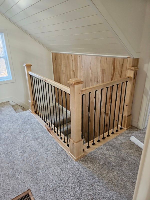Staircase with light wood railing and black spindles, carpeted floor, and wooden wall paneling in a room with angled ceiling. Staircase with light wood railing and black spindles, carpeted floor, and wooden wall paneling in a room with angled ceiling.