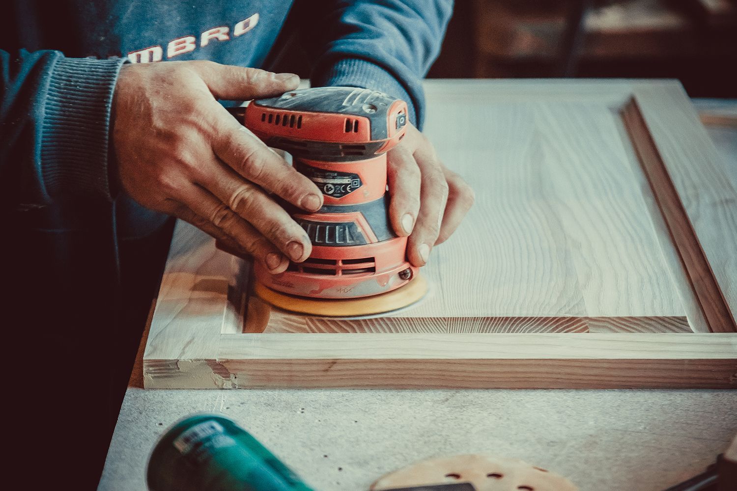 Person sanding wooden door with an orbital sander in a workshop. Person sanding wooden door with an orbital sander in a workshop.
