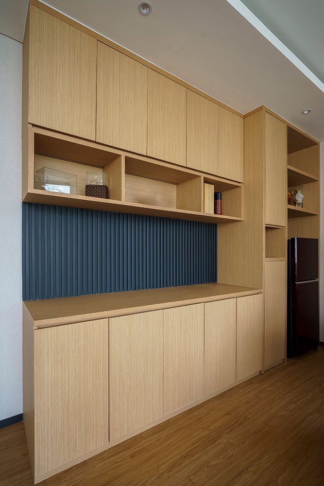 Wooden built-in storage unit with cabinets and open shelves against a blue textured wall, next to a fridge.