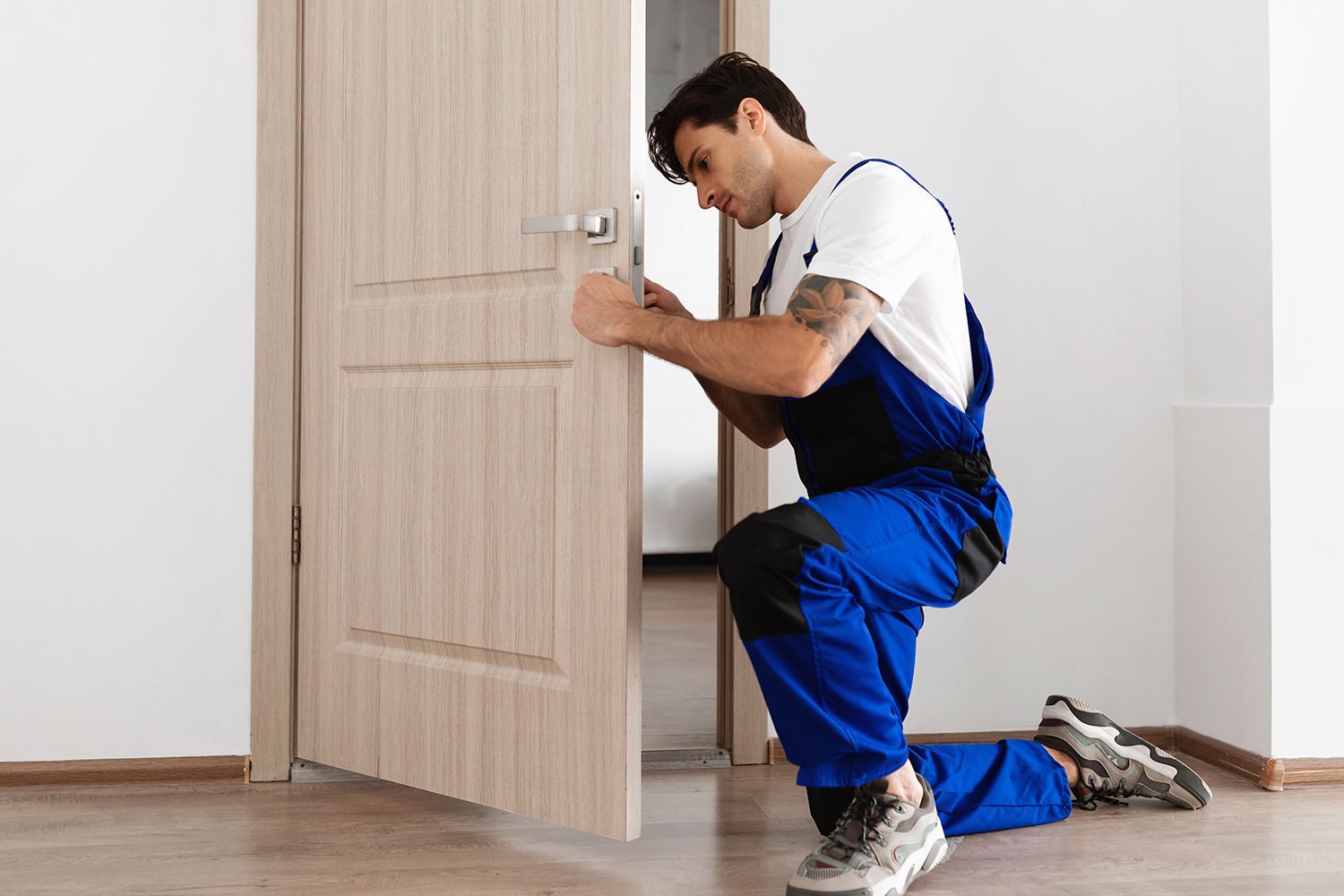 Man in blue overalls kneels, installing door handle on a beige door in a room with light wood flooring. Man in blue overalls kneels, installing door handle on a beige door in a room with light wood flooring.