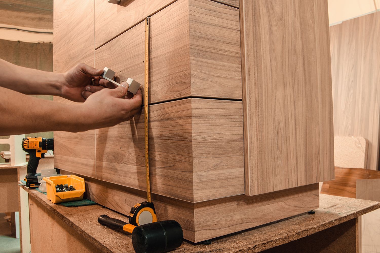 Person installing drawer pulls on a wooden cabinet; tools and tape measure visible. Person installing drawer pulls on a wooden cabinet; tools and tape measure visible.