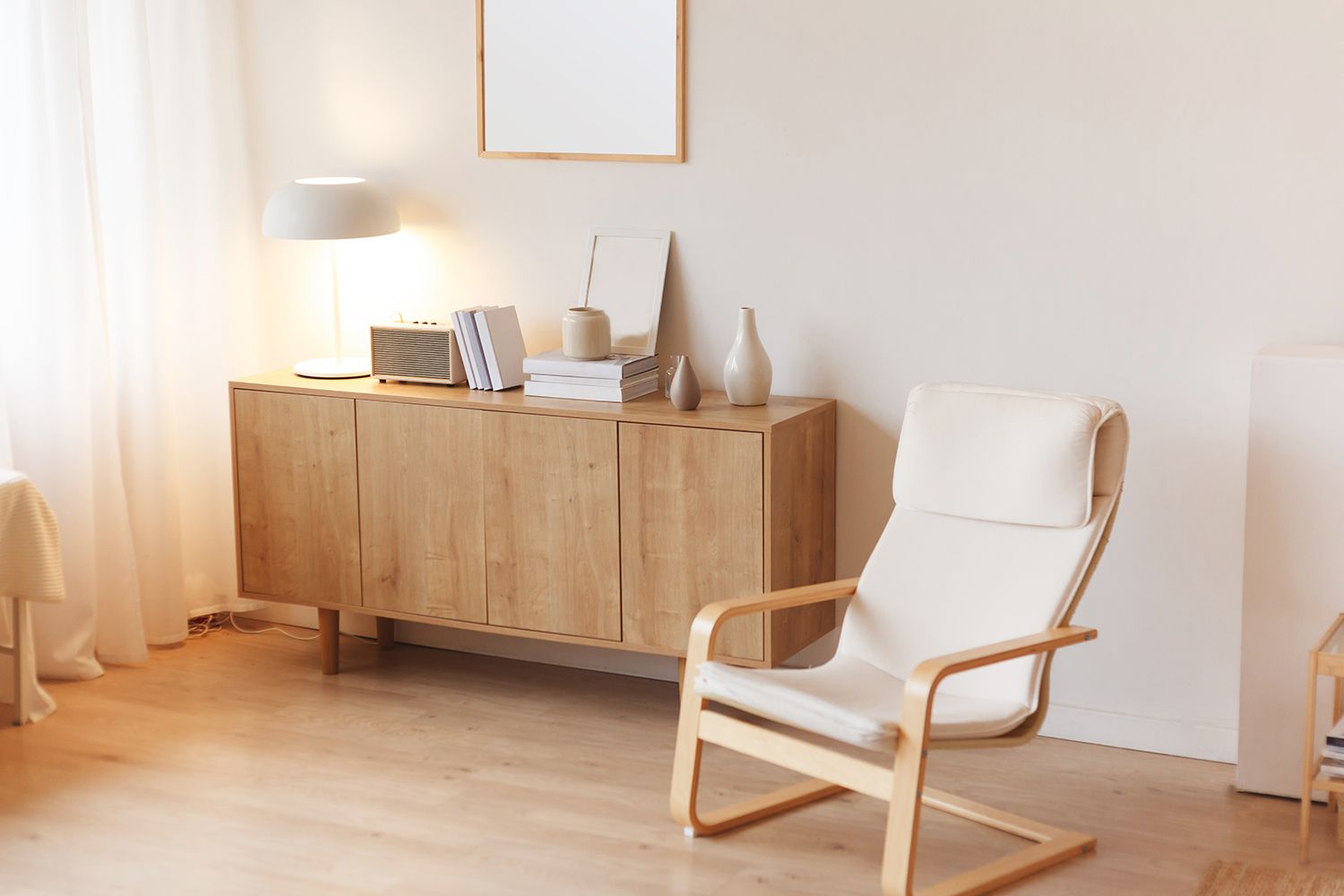 Wooden cabinet with lamp, books, and decor, beside a white armchair in a bright room. Wooden cabinet with lamp, books, and decor, beside a white armchair in a bright room.