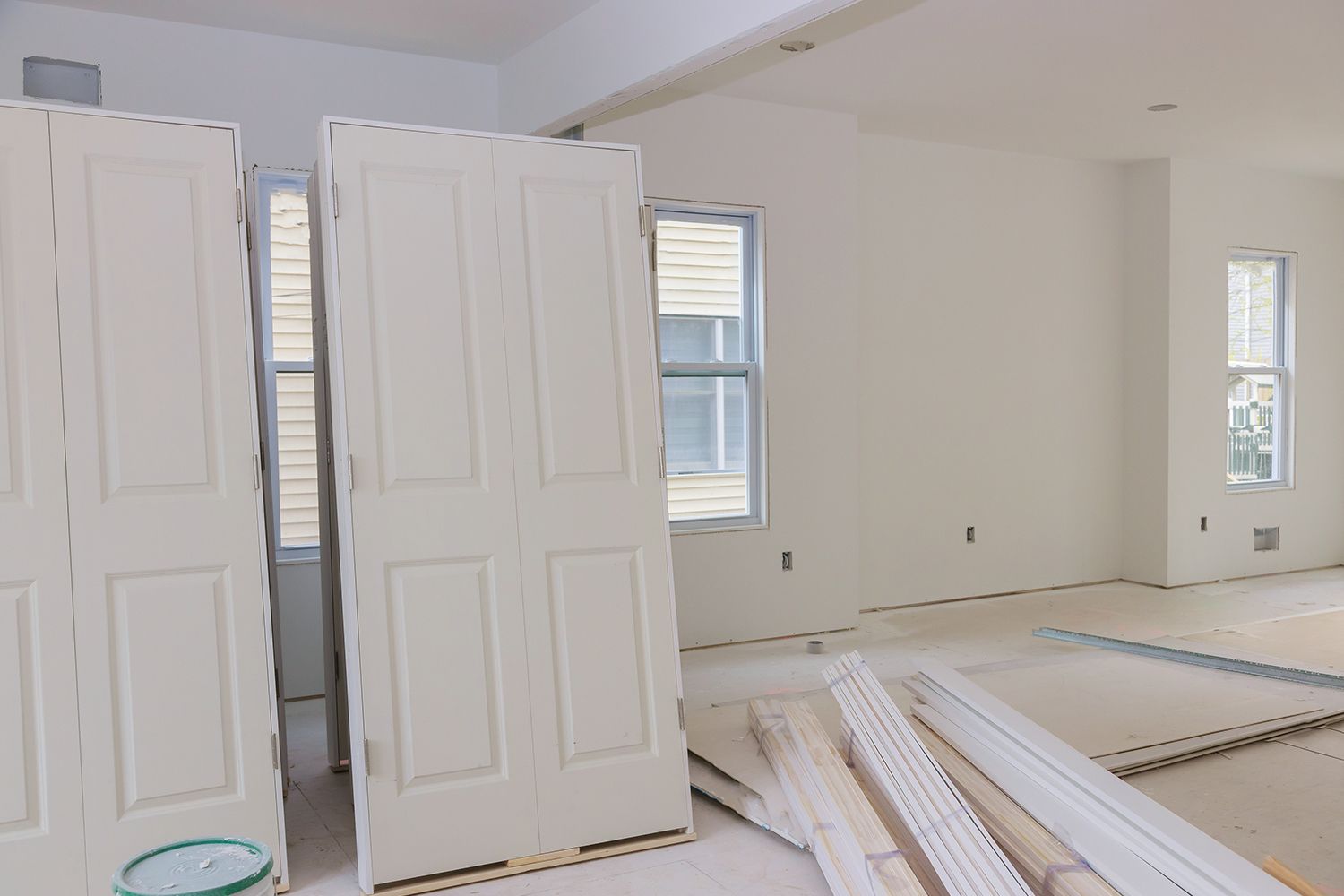 Interior of a room under construction with white walls, doors, and trim pieces. Windows and a paint bucket are also visible. Interior of a room under construction with white walls, doors, and trim pieces. Windows and a paint bucket are also visible.