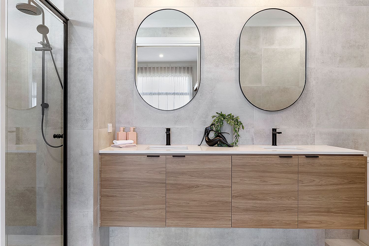 Modern bathroom with light wood vanity, two oval mirrors, black fixtures, and a shower. Modern bathroom with light wood vanity, two oval mirrors, black fixtures, and a shower.