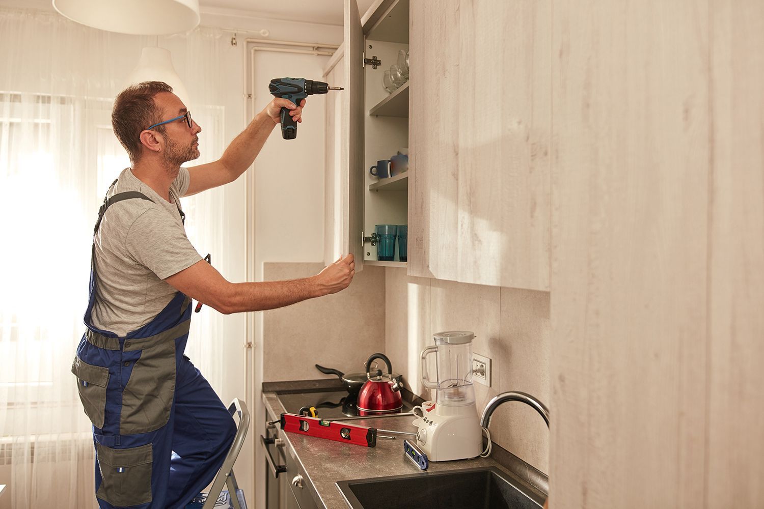 Man using a drill on a kitchen cabinet, standing on a step stool. Man using a drill on a kitchen cabinet, standing on a step stool.