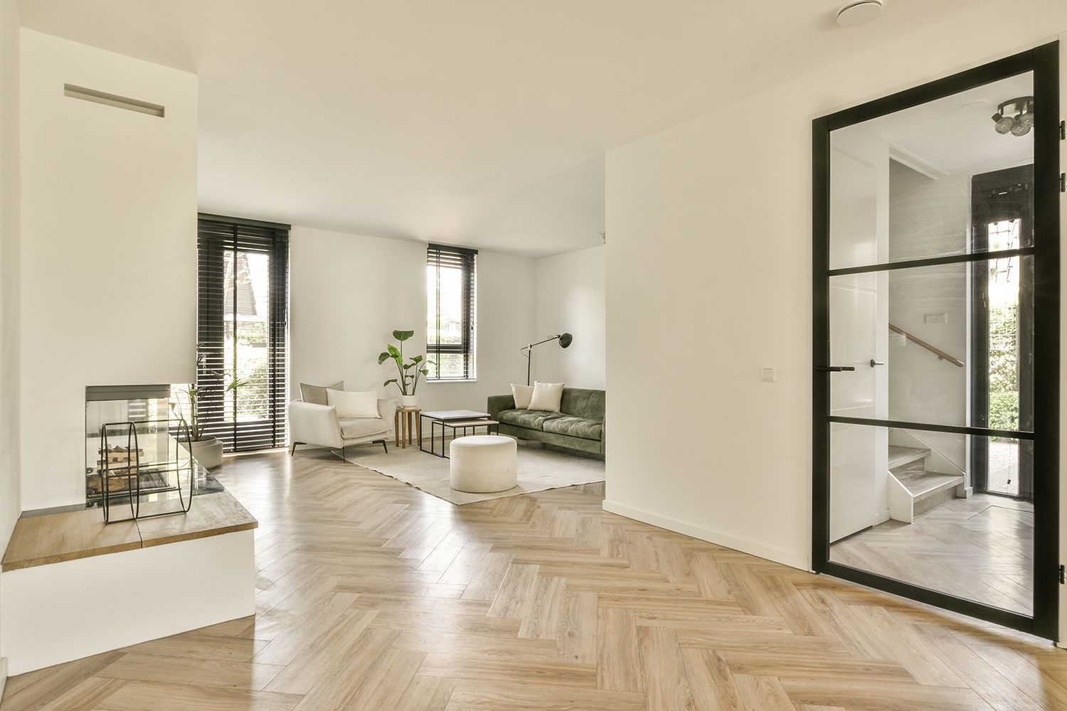Living room with herringbone wood floors, a green sofa, and black-framed glass door to a staircase. Living room with herringbone wood floors, a green sofa, and black-framed glass door to a staircase.
