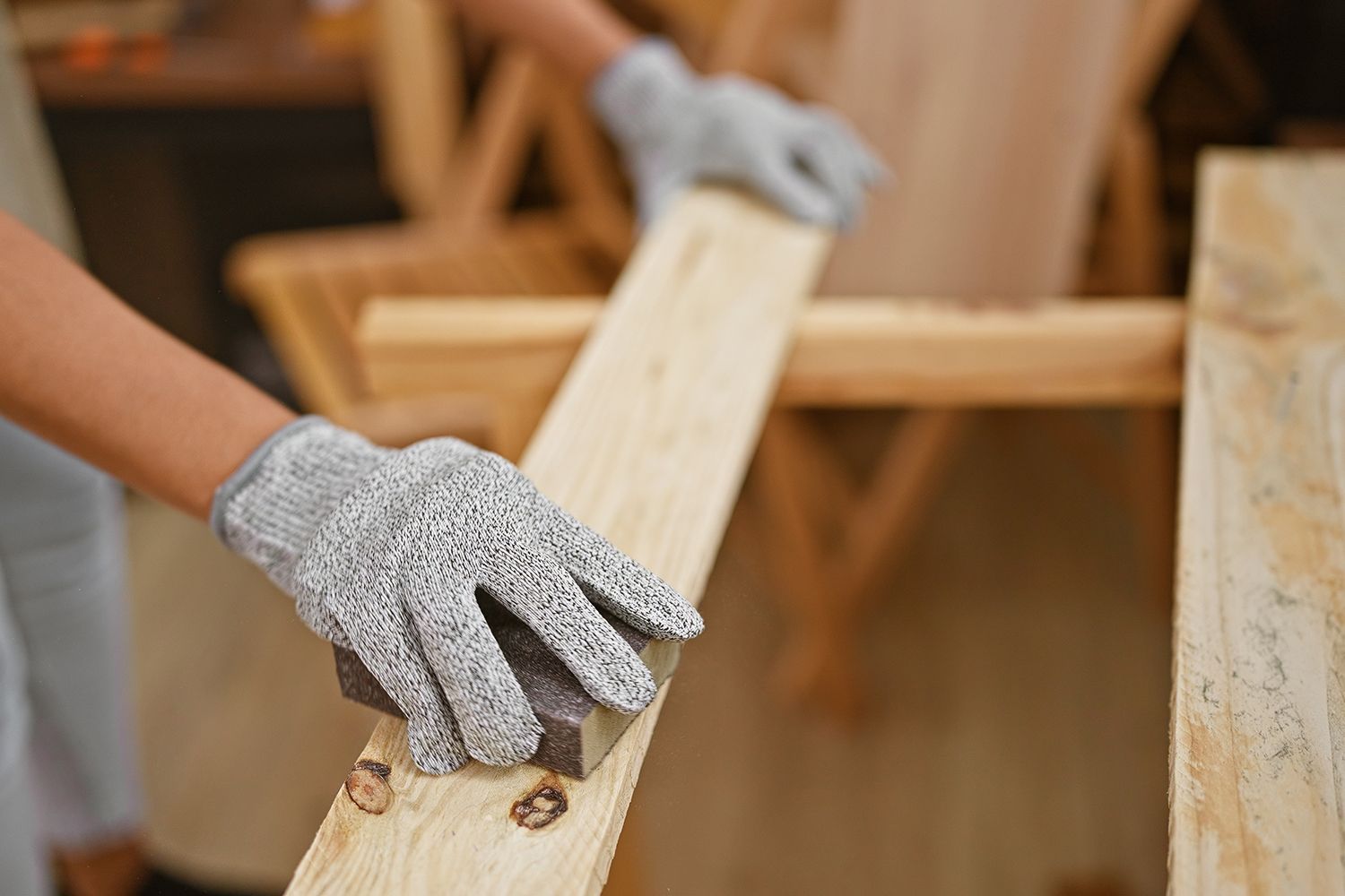 Person sanding a wooden plank with a sanding block, wearing gray gloves, in a workshop.
