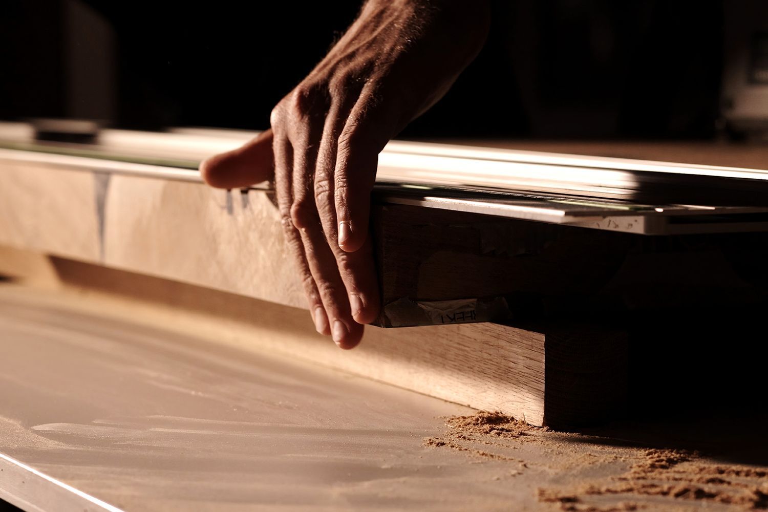 Hand of a person guiding wood along a saw blade, sawdust visible, dimly lit workshop.