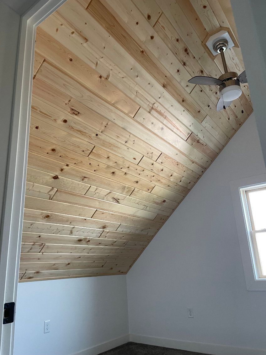 Angled attic room with light wood plank ceiling, white walls, and a ceiling fan.