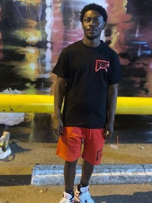 A young man wearing a black t-shirt and red shorts is standing in front of a wall.