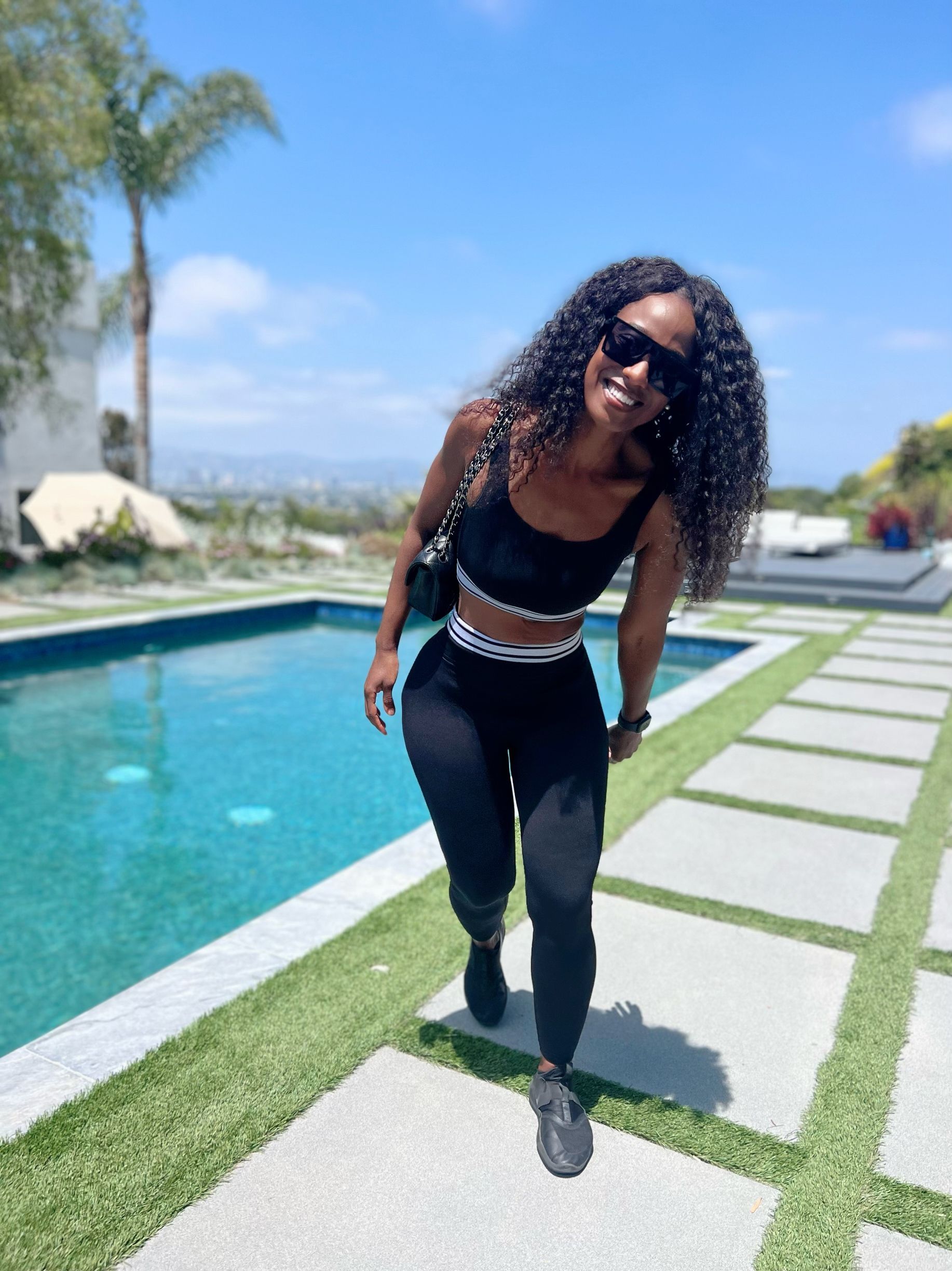 Woman standing by the pool with sunglasses on, smiling at the camera. 