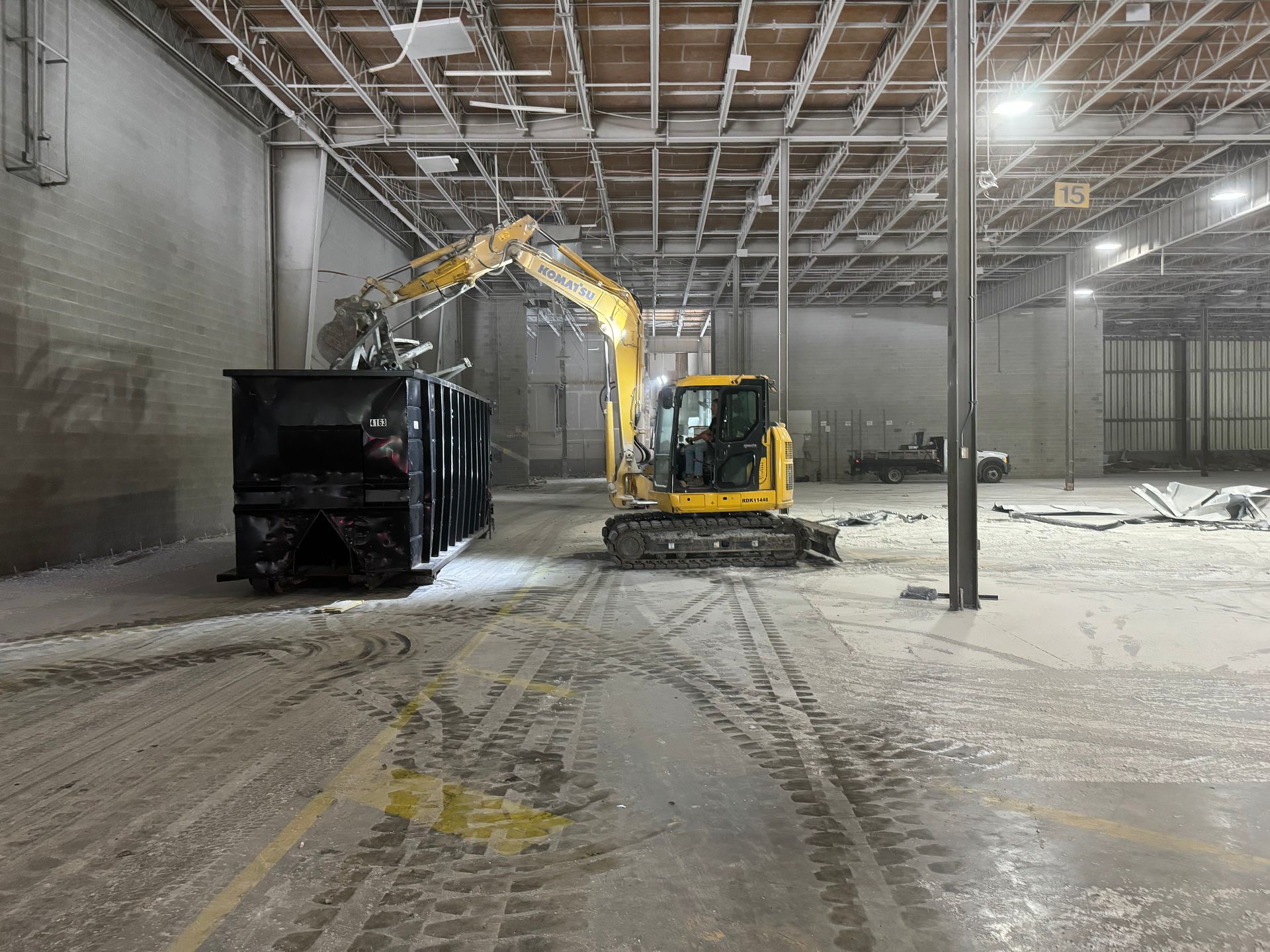 A yellow excavator is loading a dumpster into a large warehouse.