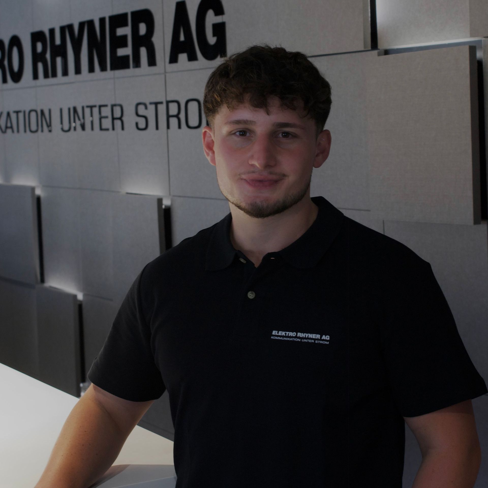 Ein Mann in einem schwarzen Hemd steht vor einem Schild mit der Aufschrift „Ro Rhyner AG“