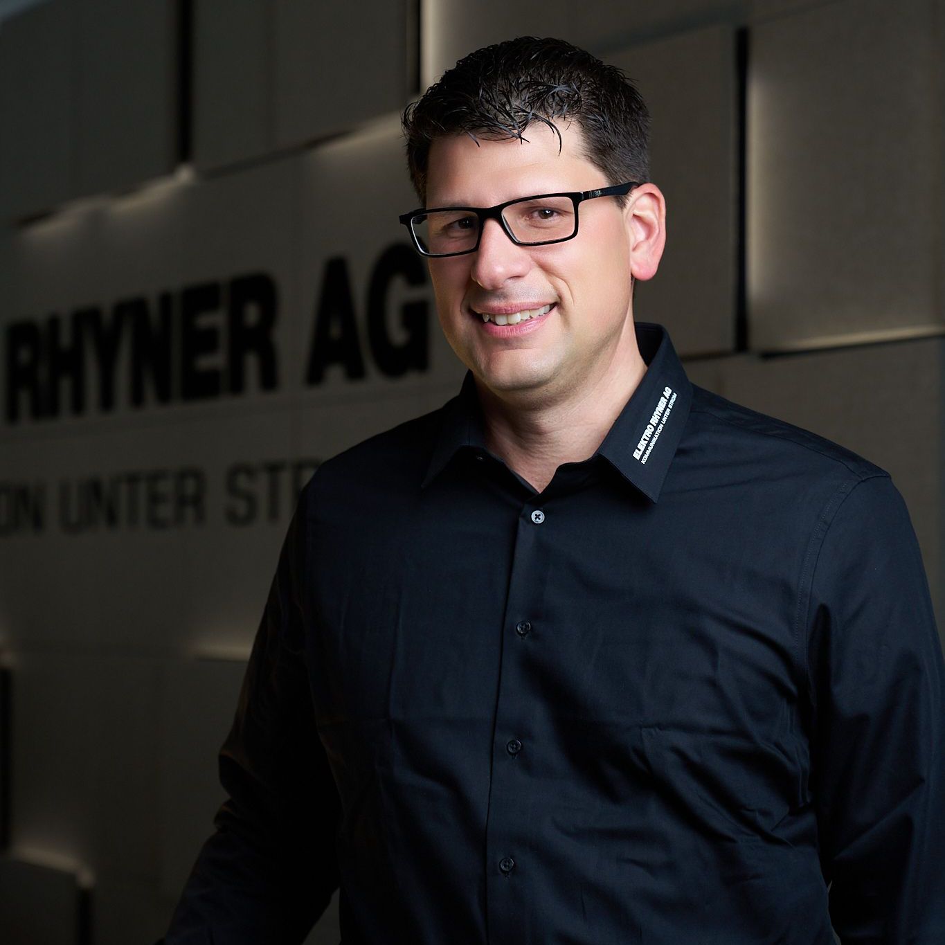 Ein Mann mit Brille und schwarzem Hemd steht vor einem Schild mit der Aufschrift „Rhyner AG“