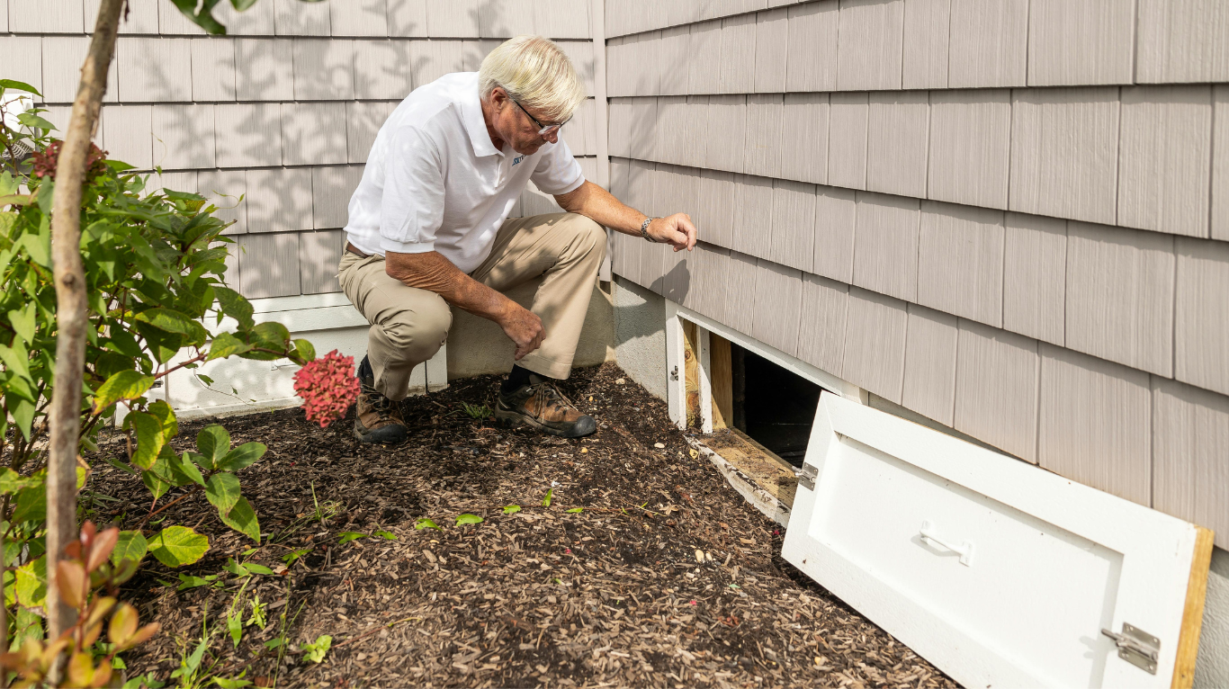 Homeowner inspecting a crawl space access opening along the exterior foundation of a house.