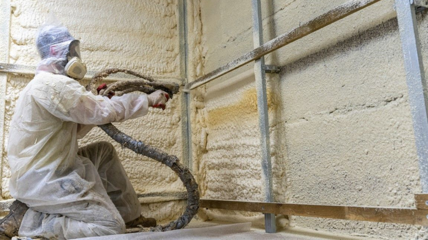 Installer spraying foam insulation onto metal wall panels inside a pole barn structure.