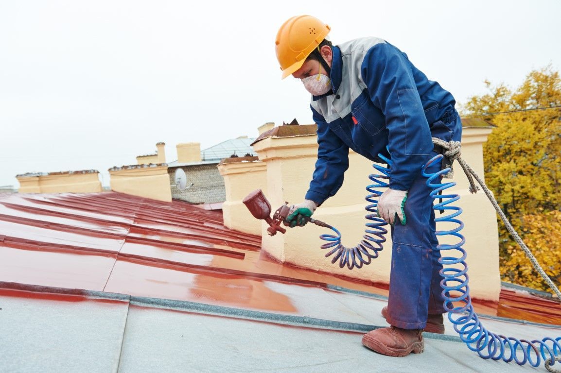Man in safety gear spray-painting a red roof with a spray gun, outdoors.