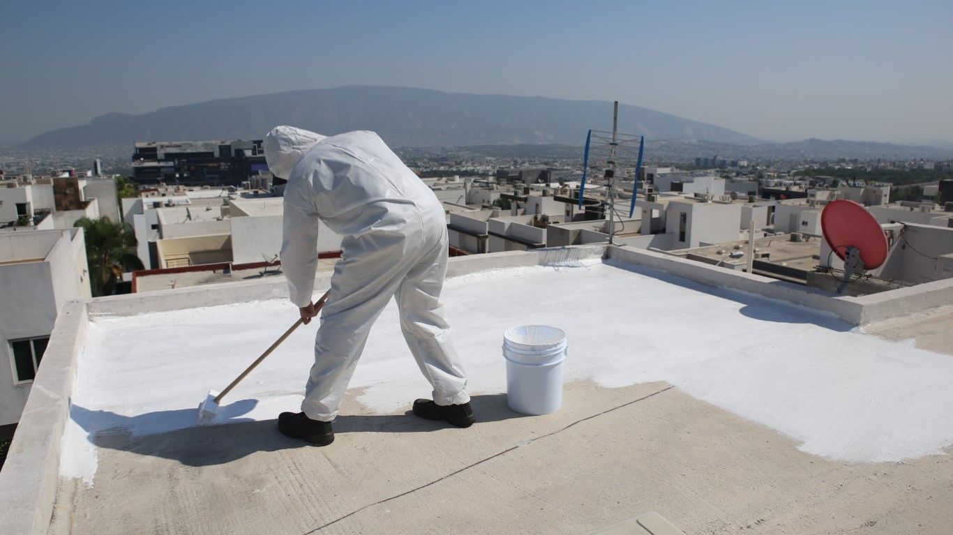 Person in white protective suit painting a white coating on a rooftop, sunny day.