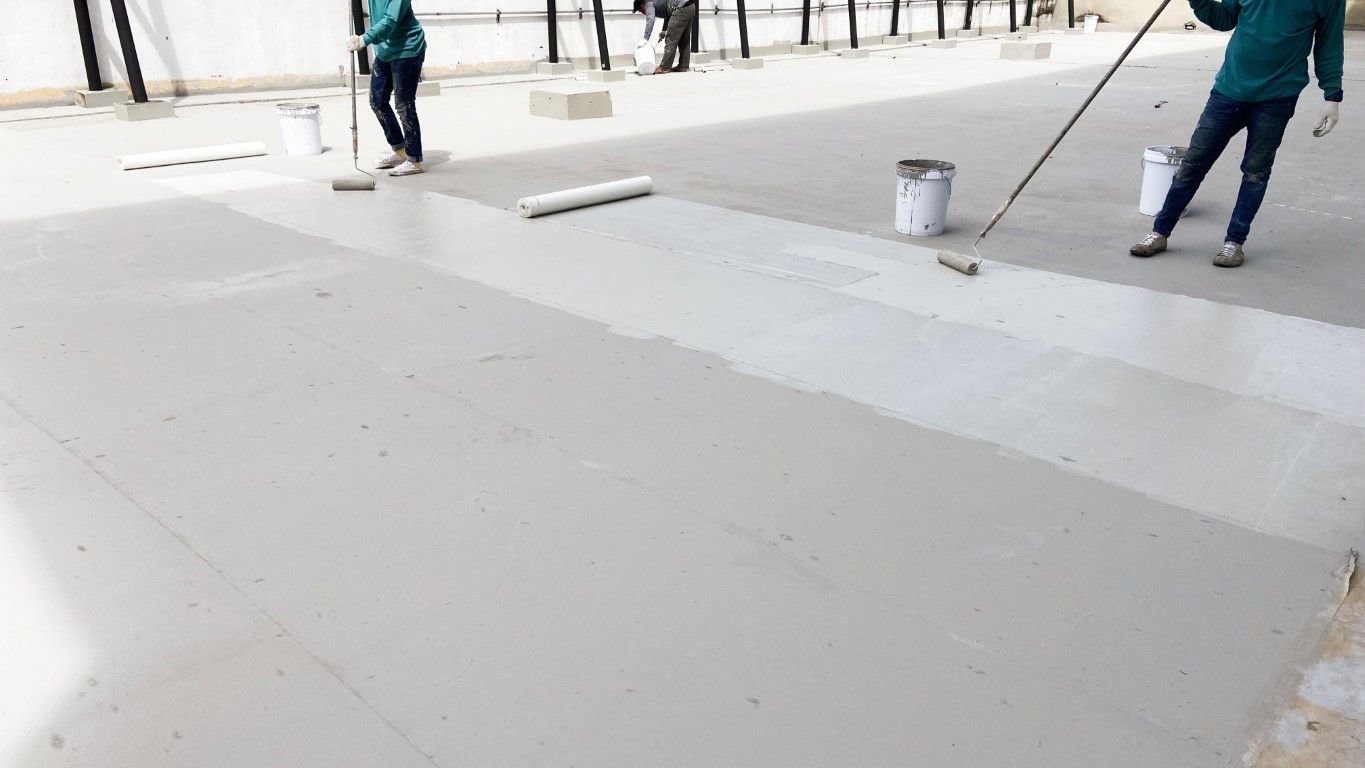 Workers applying white coating to a concrete floor with rollers, buckets of paint nearby, outdoors.