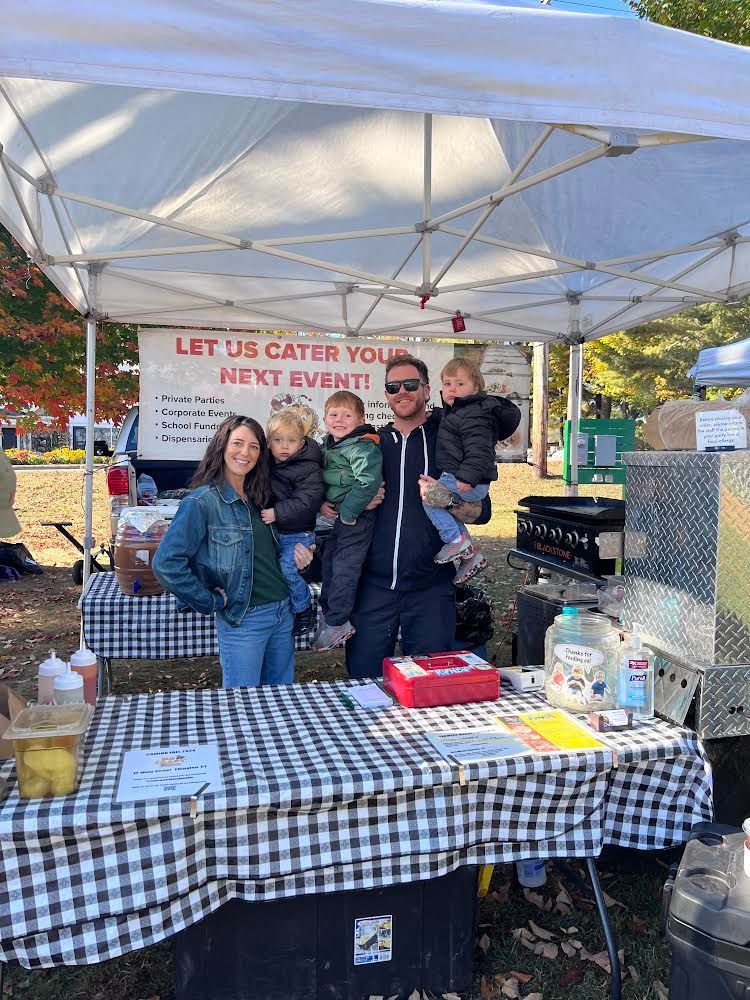 Owners of No Coast Tacquera stands behind a catering booth with a white canopy and checkered tablecloth at an outdoor event.