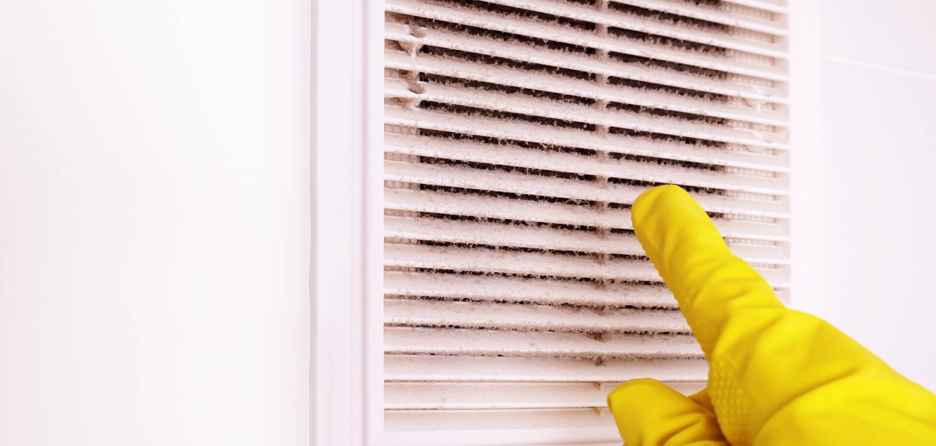 A hand wearing a yellow rubber glove pointing at a dirty air vent