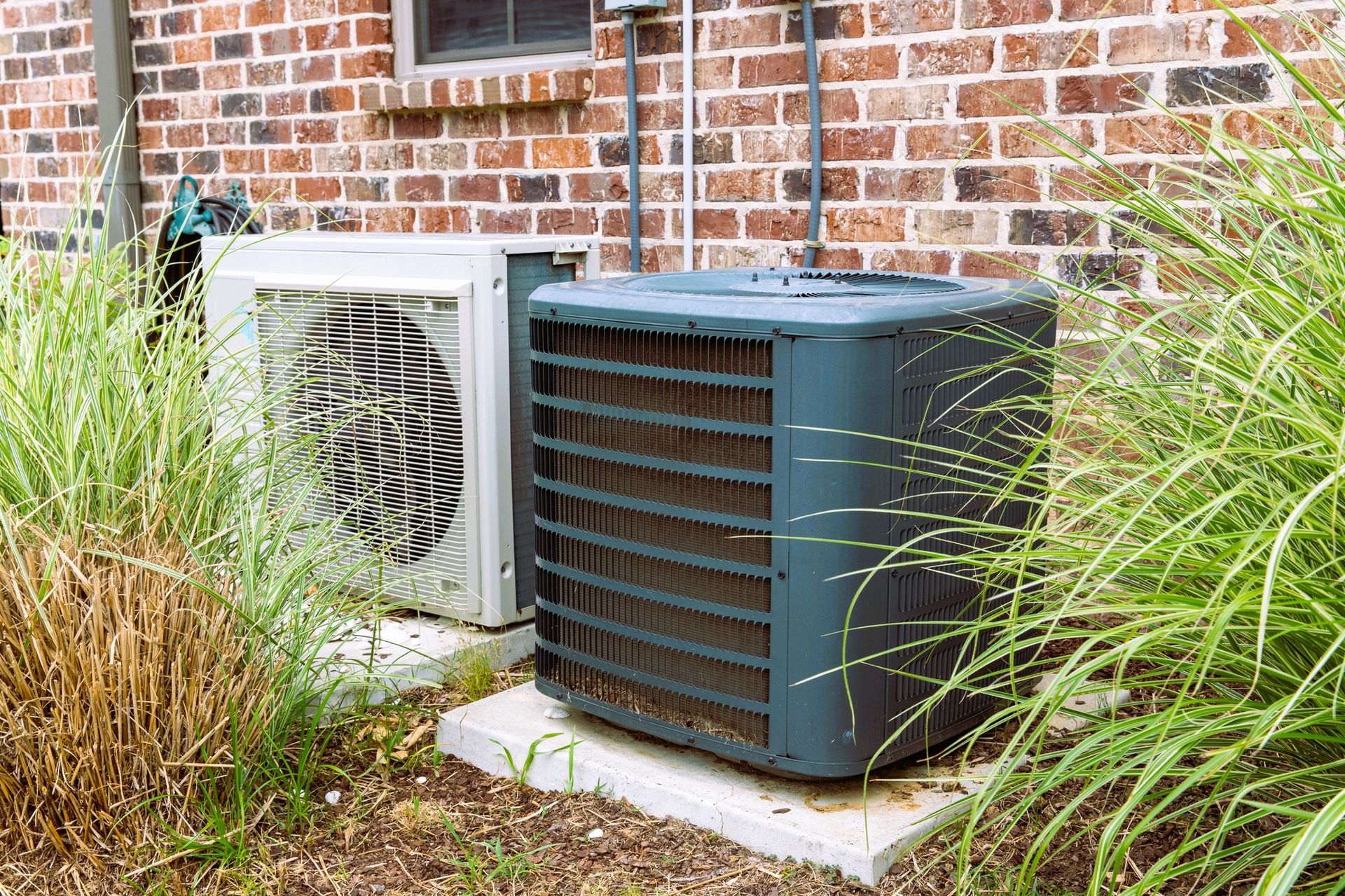 HVAC units outside a brick house surrounded by overgrown grass.