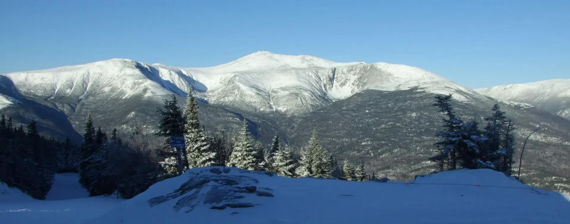 A snowy mountain range with trees in the foreground and a blue sky in the background.