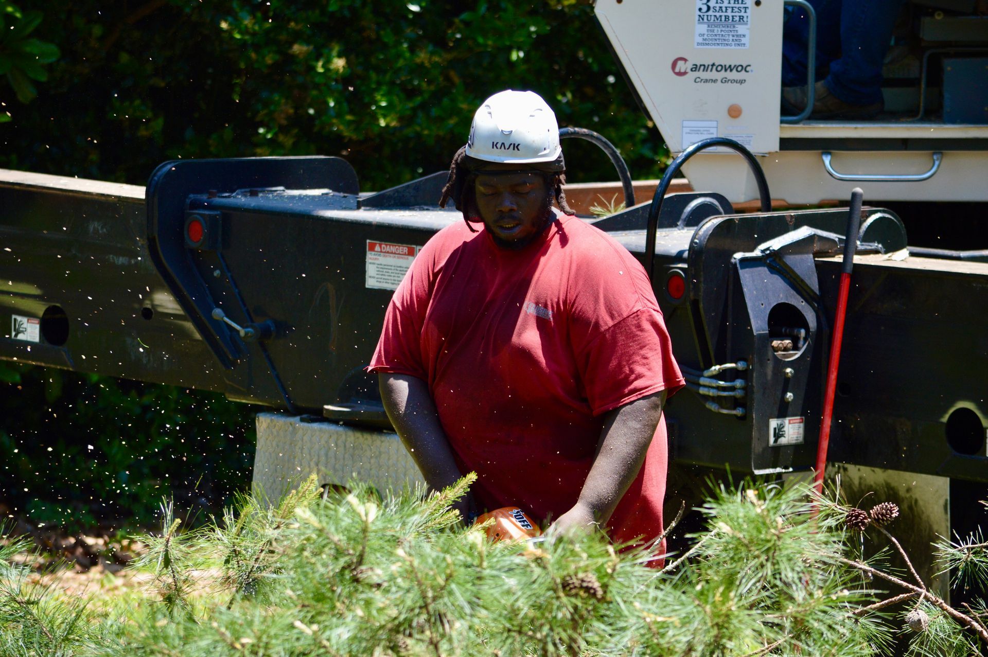 Person in red shirt and hard hat cutting brush with a chainsaw near a wood chipper.