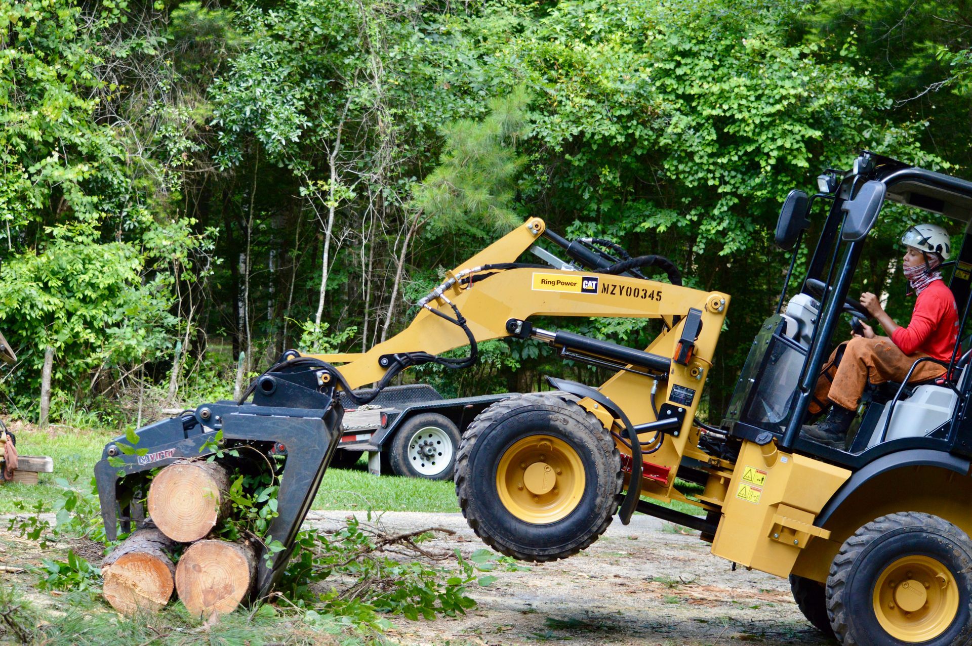 Yellow forklift with a grapple attachment lifting logs in a wooded area.