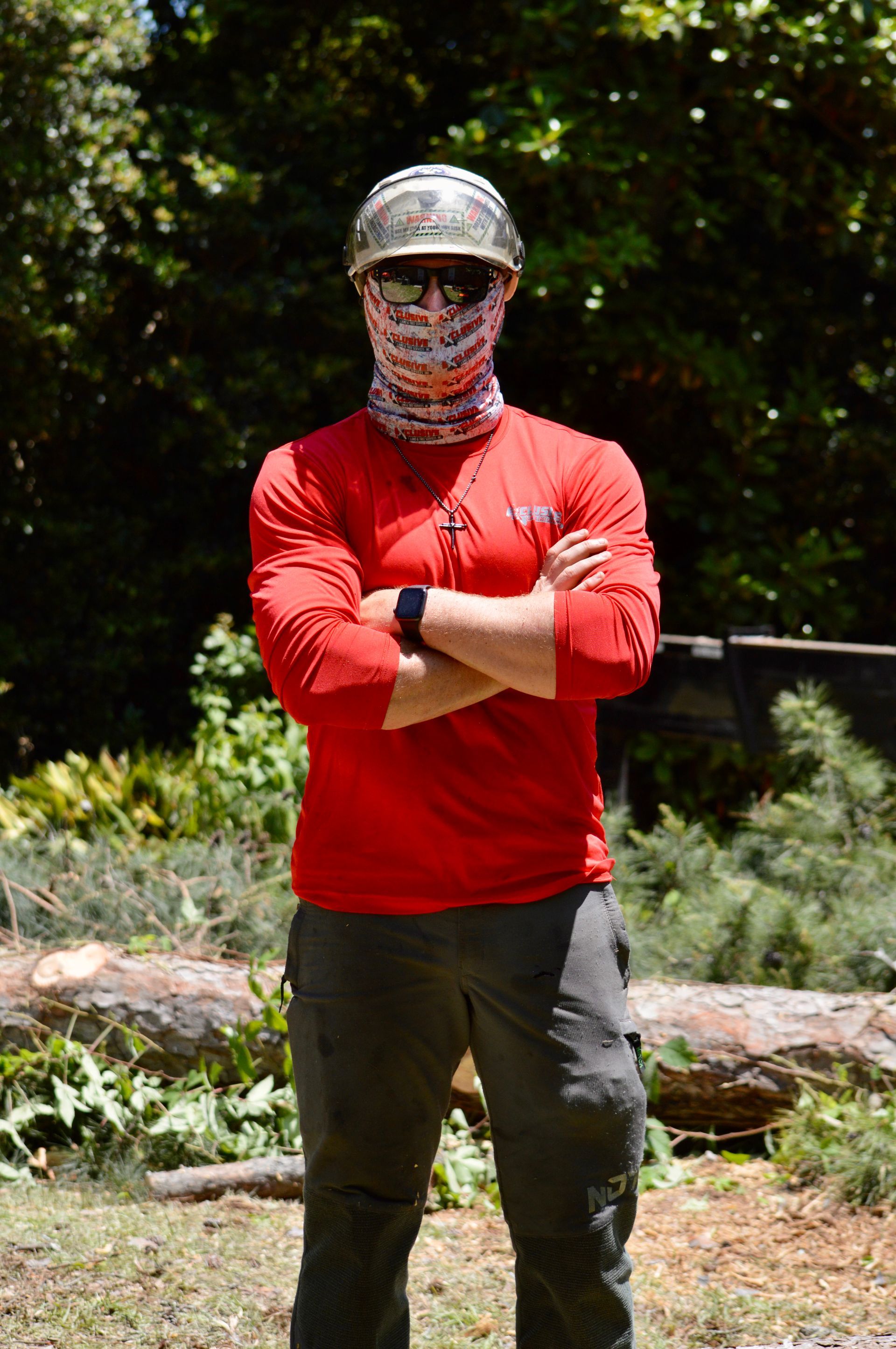 Man with arms crossed, wearing a helmet, red shirt, and face covering, standing in a wooded area.