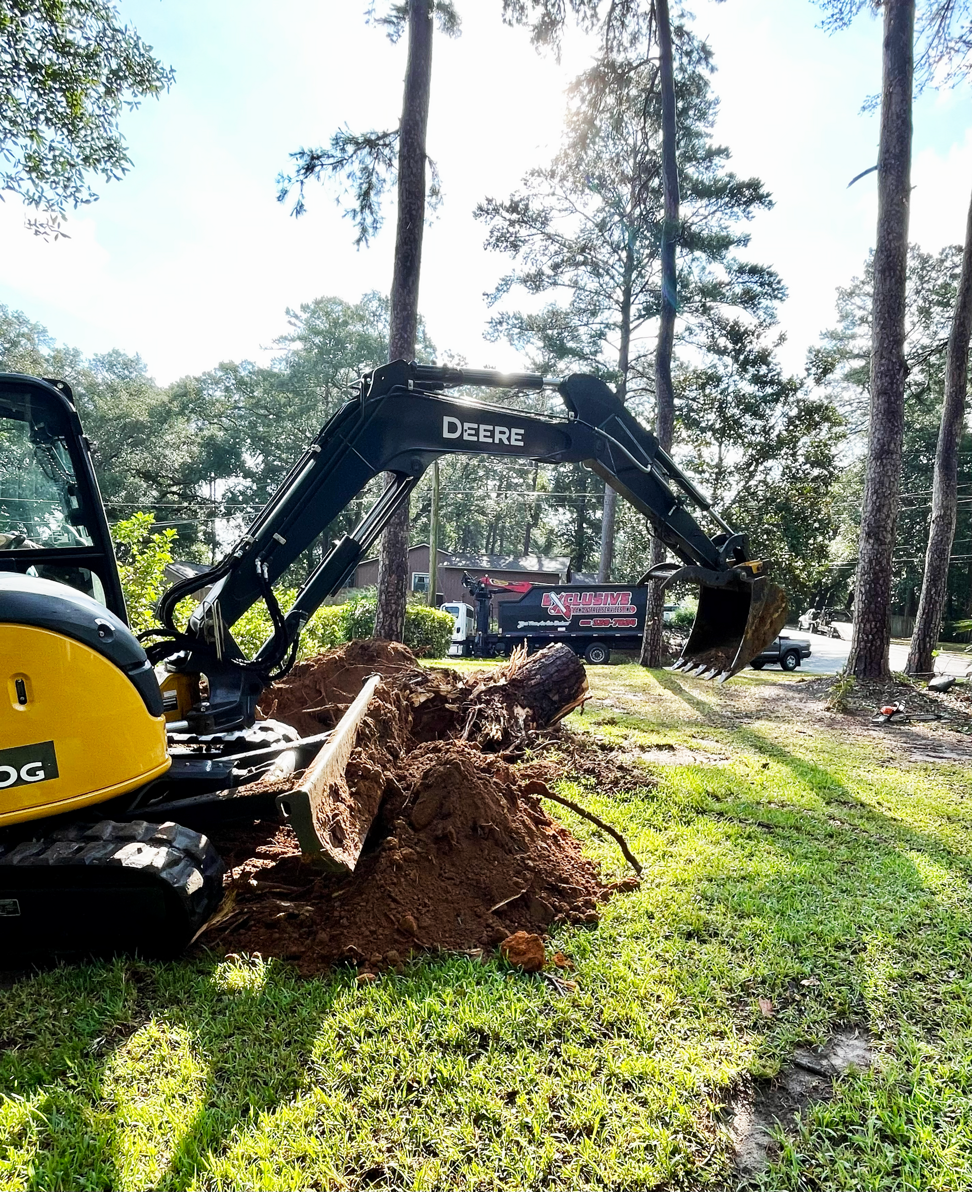 A mini excavator digging a hole in grassy area, with trees and a truck in the background.