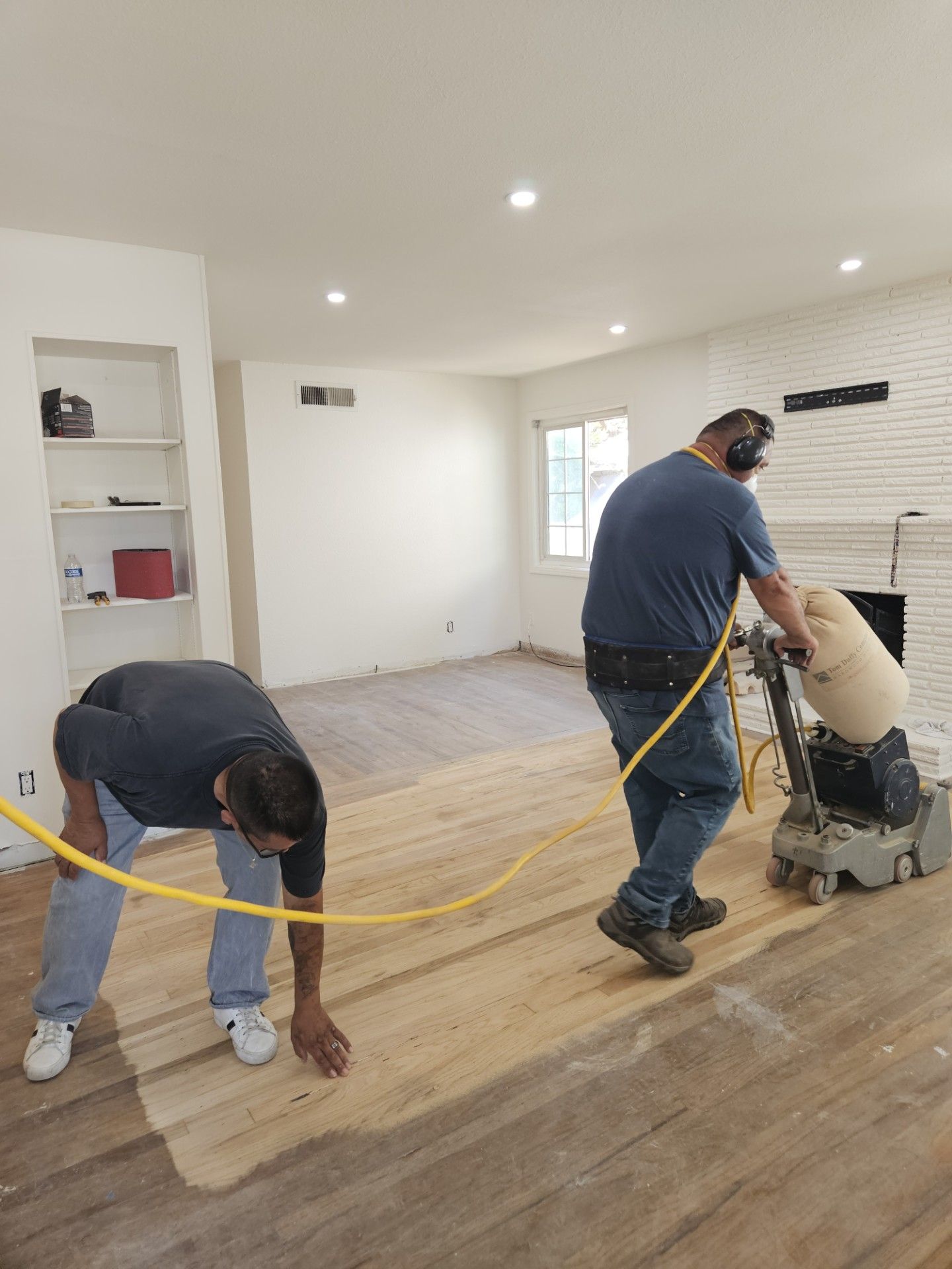 Two men are sanding a wooden floor in a living room.