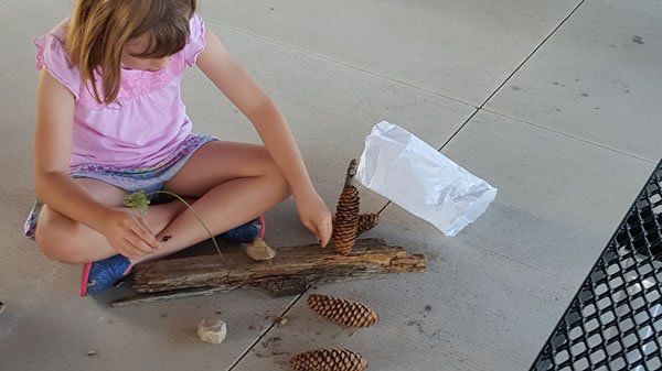 A little girl is sitting on the ground making a boat out of pine cones.