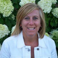 Amy Hawkins in a white jacket is smiling in front of a bush of flowers.