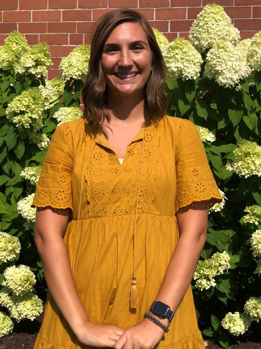 Lynn in a yellow dress is standing in front of a bush of flowers.