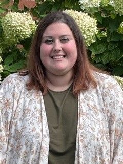 Sarah is smiling in front of a bush of flowers.