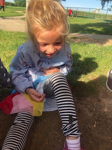 A little girl wearing striped leggings is sitting on the ground in a park.
