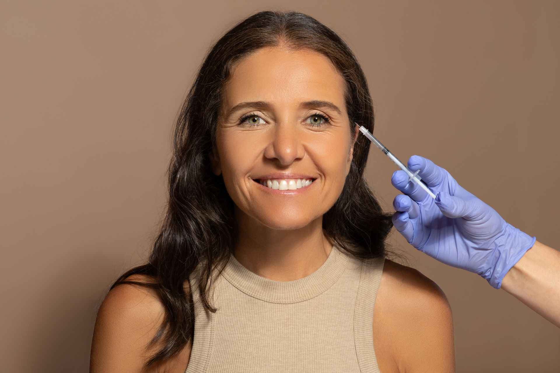 Woman smiles as a gloved hand holds a syringe near her eyebrow; brown background.