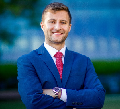 A man in a blue suit and red tie is standing with his arms crossed and smiling.