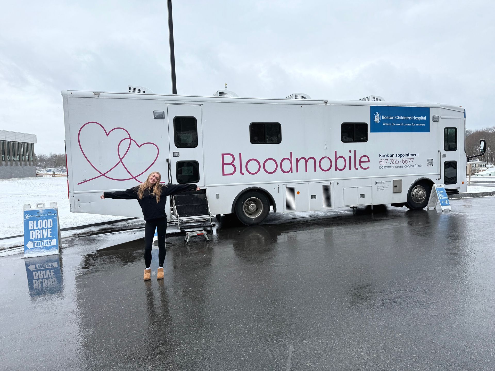A woman is standing in front of a bloodmobile in a parking lot.