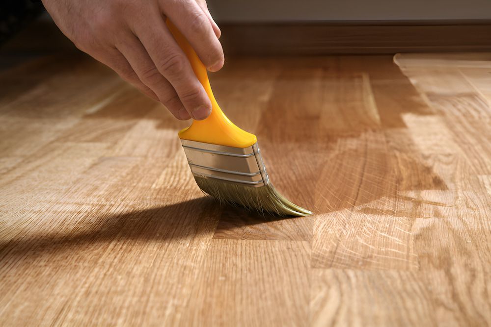 A Person Is Painting A Wooden Floor With A Brush — Kyall Chalker Floor Sanding & Polishing In Taree, NSW