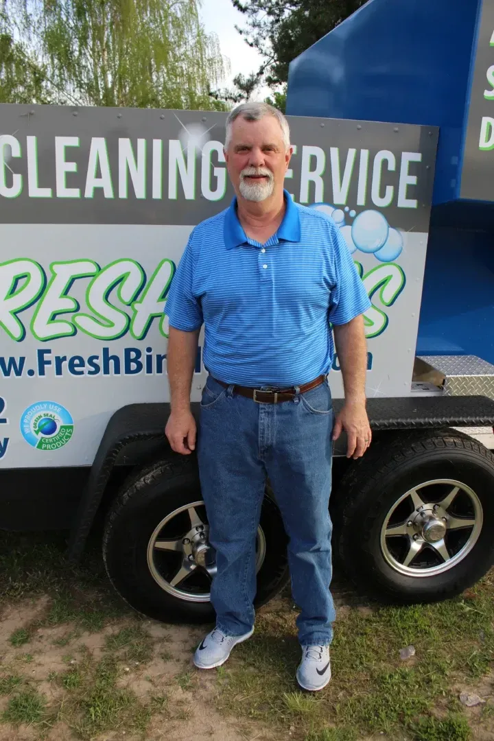 Man standing in front of a trailer for Fresh Bird Cleaning Service wearing a blue shirt and jeans.