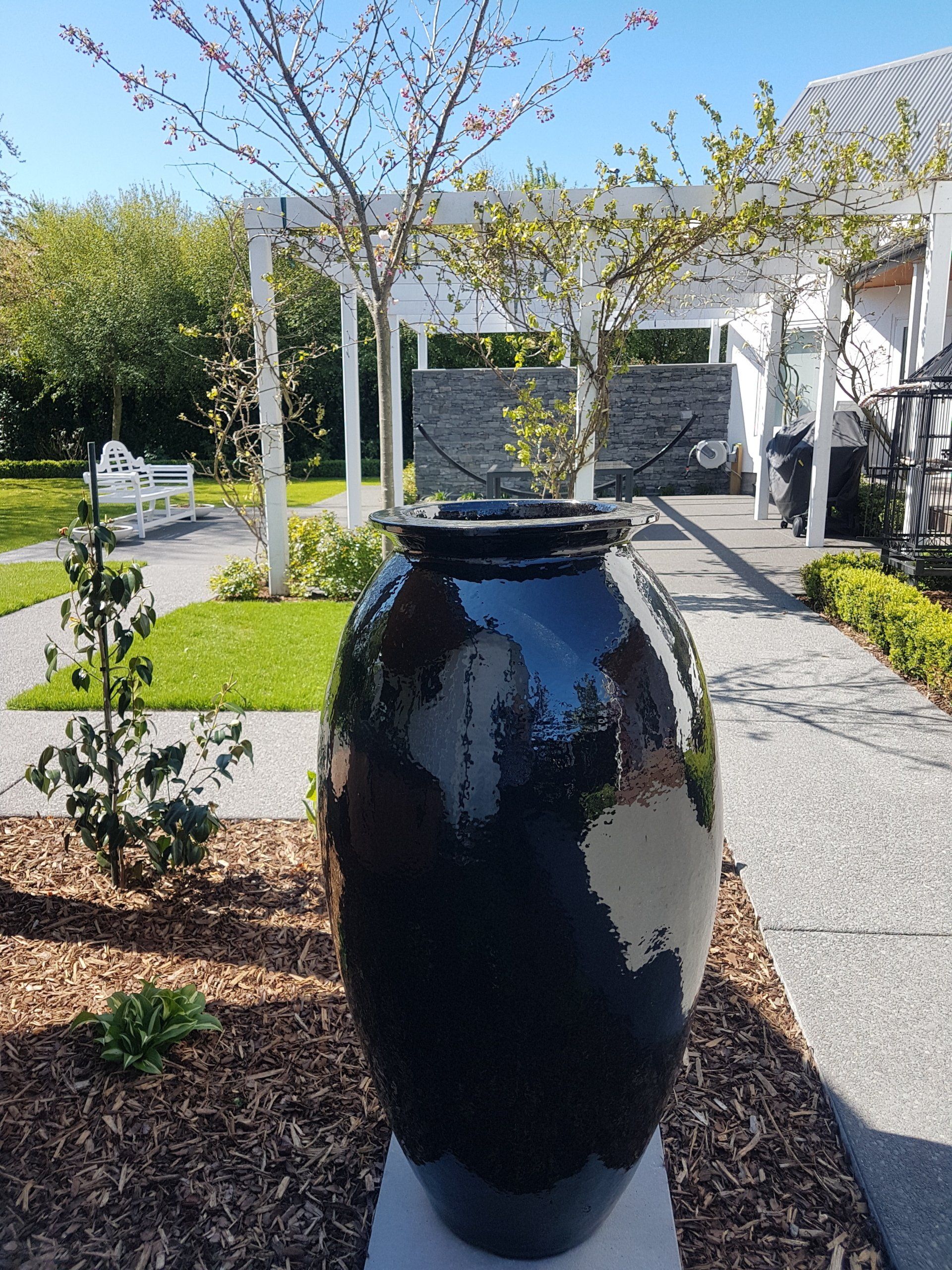 A large black vase sits on a pedestal in a garden