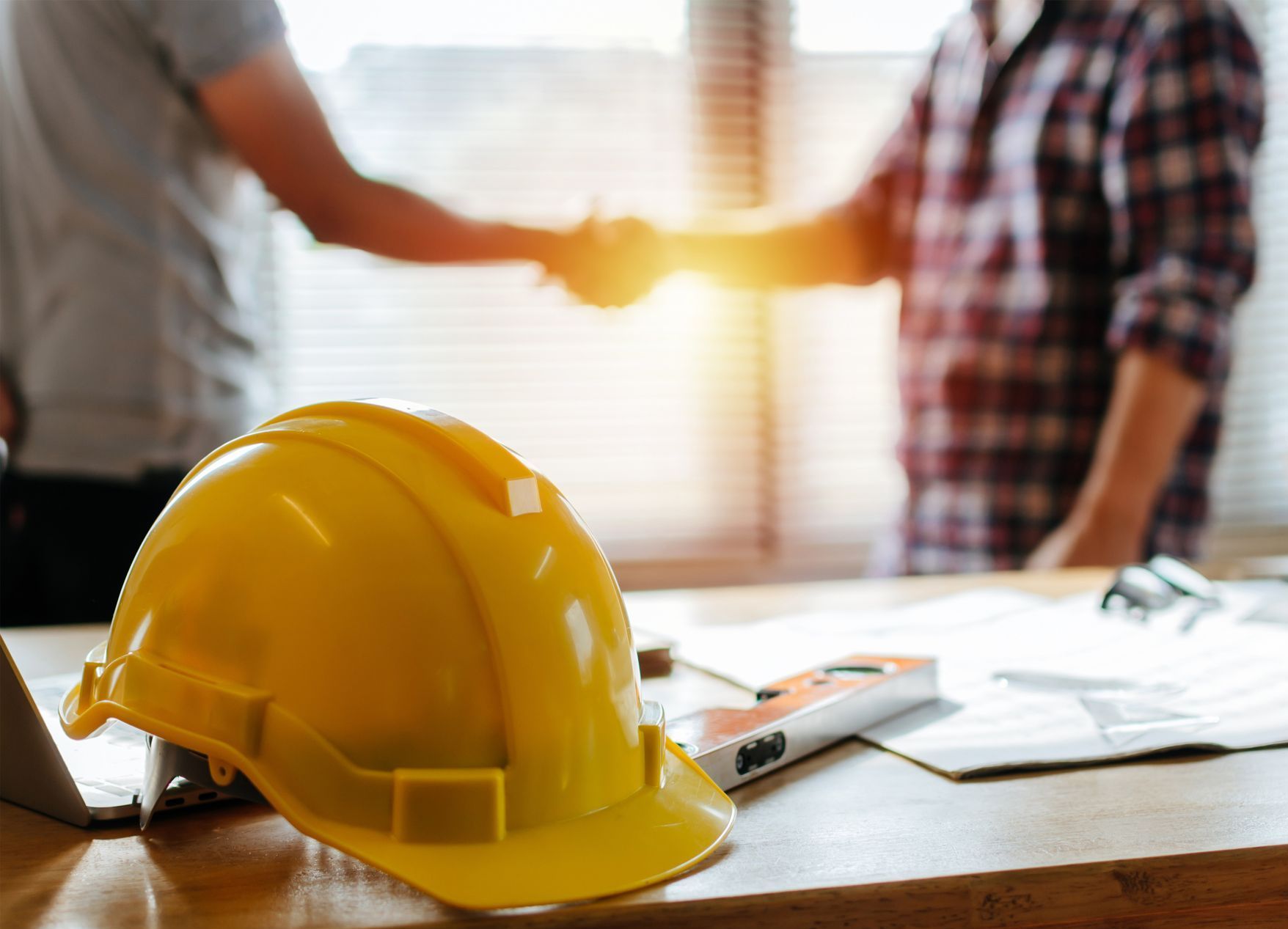 A yellow hard hat is sitting on a wooden table next to two men shaking hands.