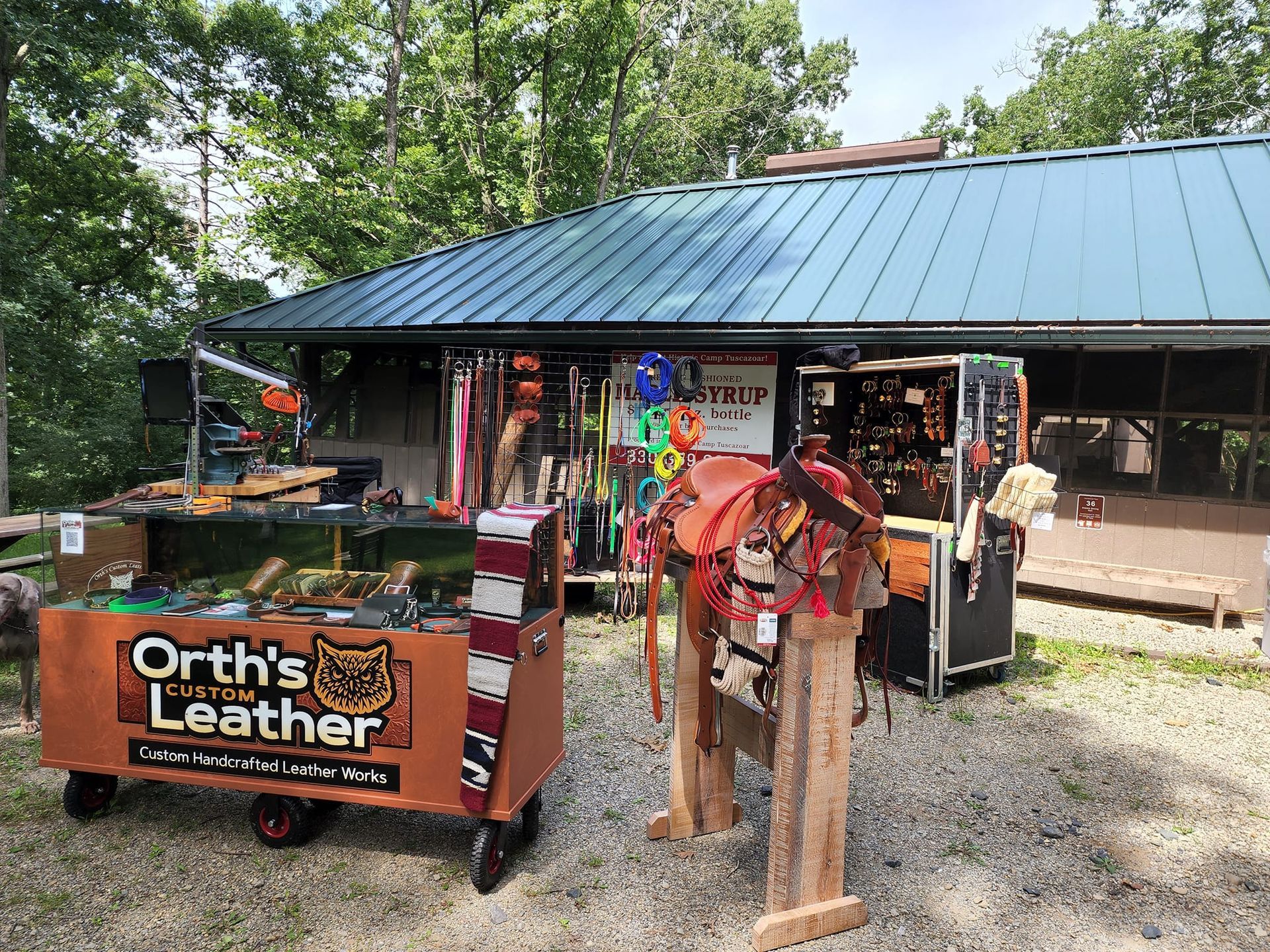 Outdoor leather craft booth with wood signs, hanging goods, and a metal-roofed shop in a wooded setting