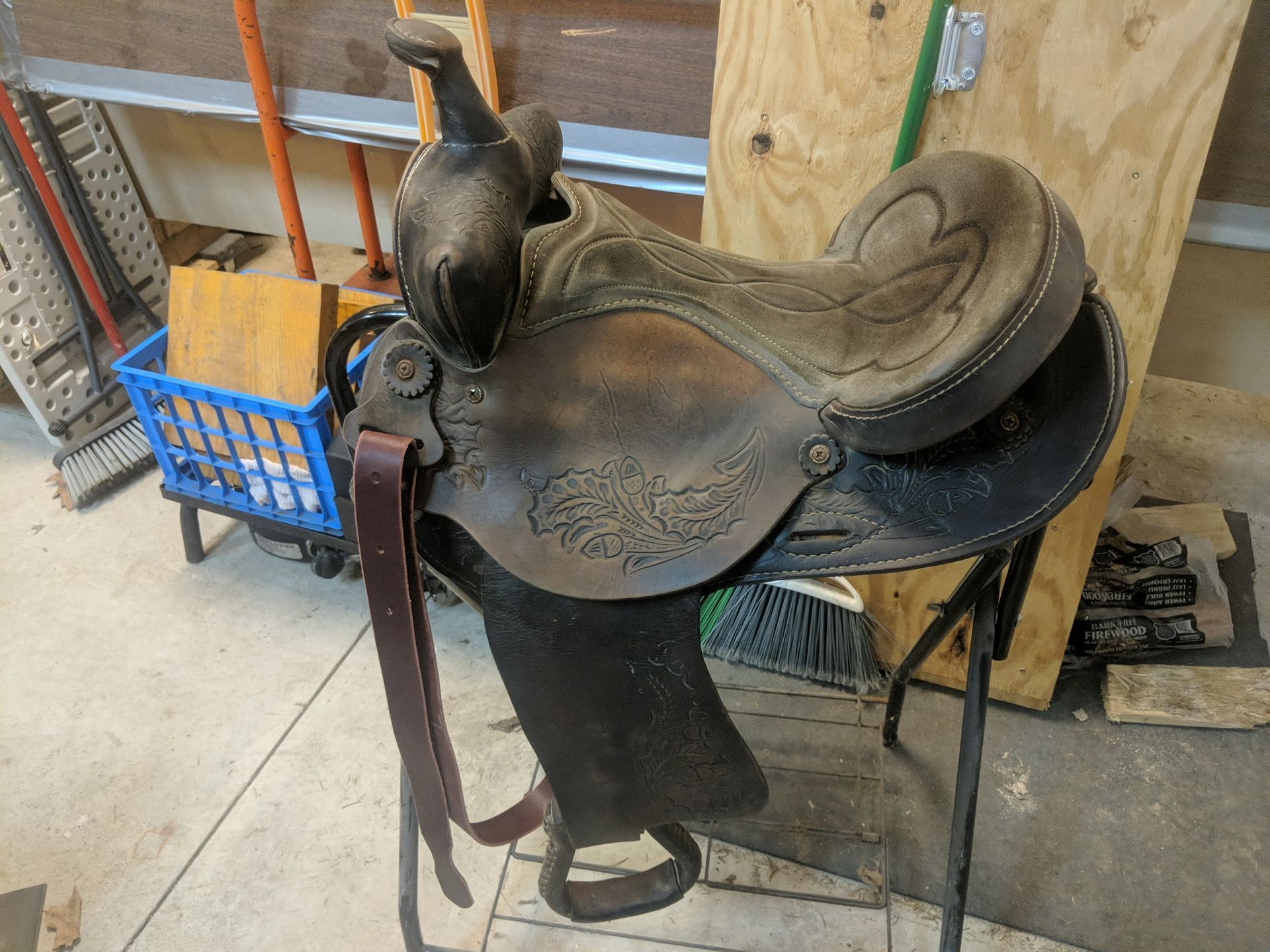 Old leather saddle on a stand in a workshop, with a blue basket and wooden boards nearby.